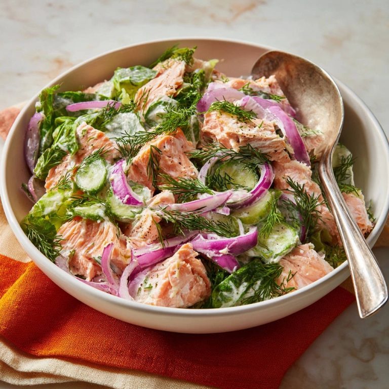 Creamy dill salmon salad served in a white bowl with a spoon, featuring flaky salmon, cucumbers, and red onion.
