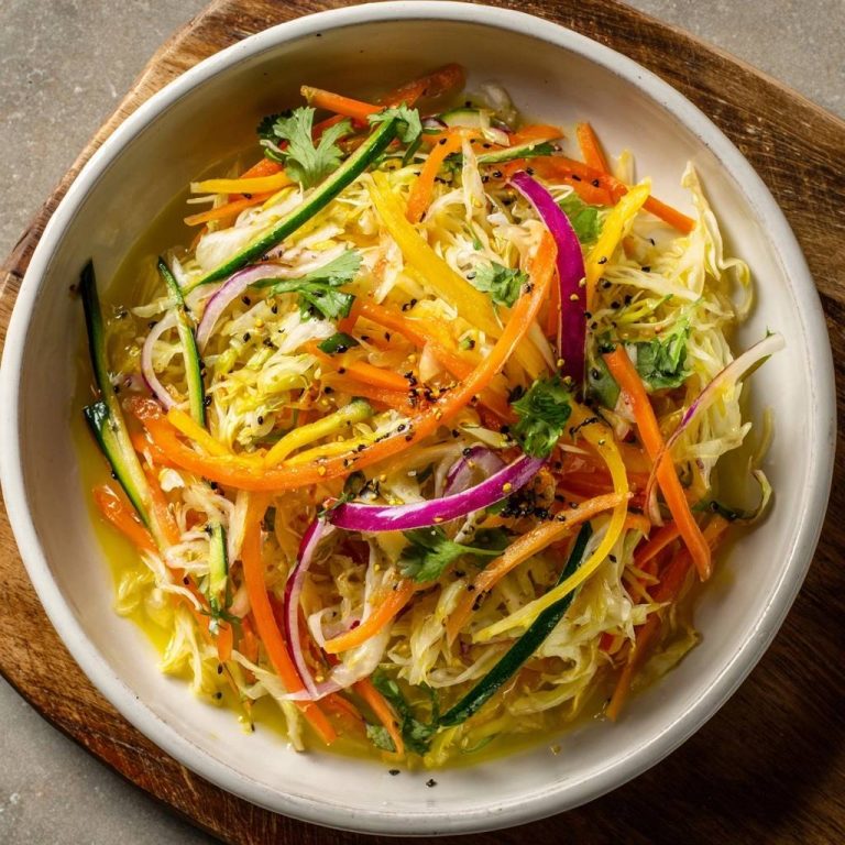 Rainbow veggie slaw in a white bowl, featuring shredded cabbage, carrots, bell peppers, red onion, and cucumber, sprinkled with seeds.