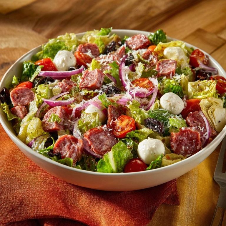 Chopped antipasto salad served in a large white bowl, featuring salami, mozzarella, tomatoes, olives, and red onion.